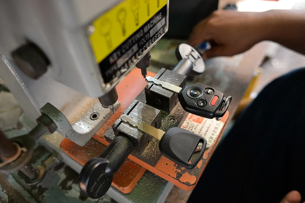 A car key with lock, unlock and panic buttons clamped in a key cutting machine beside a new blank being cut to match it.