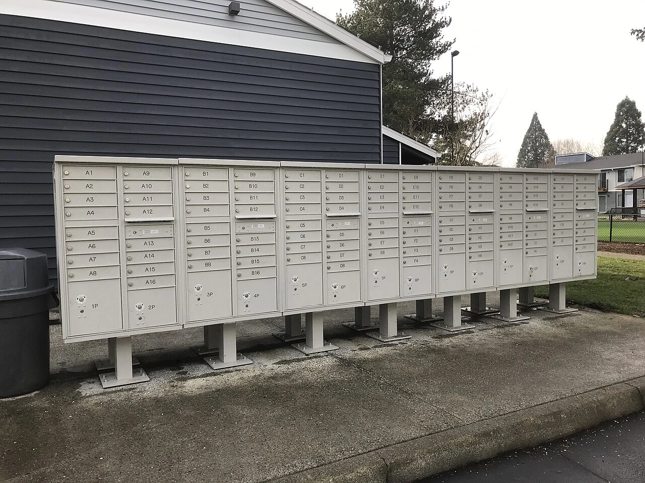 A long row of tan cluster mailboxes on posts at an apartment complex, with lettered doors and larger parcel locker doors at the bottom.