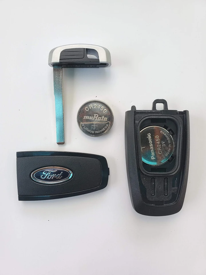 A Ford smart key taken apart, showing the two halves of the shell, the round CR2450 coin battery and the metal emergency key blade.