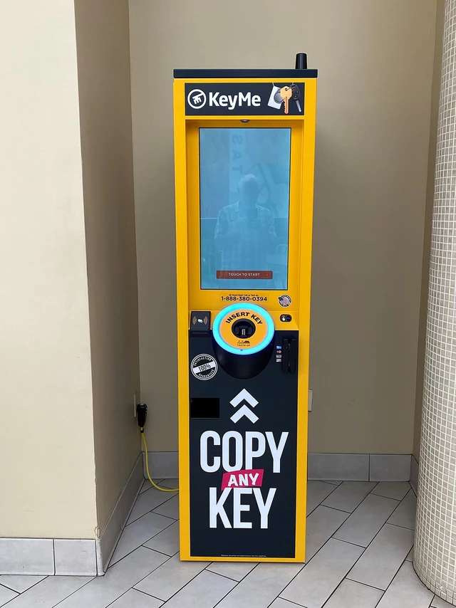 A yellow KeyMe self service key copying kiosk with a touch screen and a key slot, standing against a wall in a shopping mall.